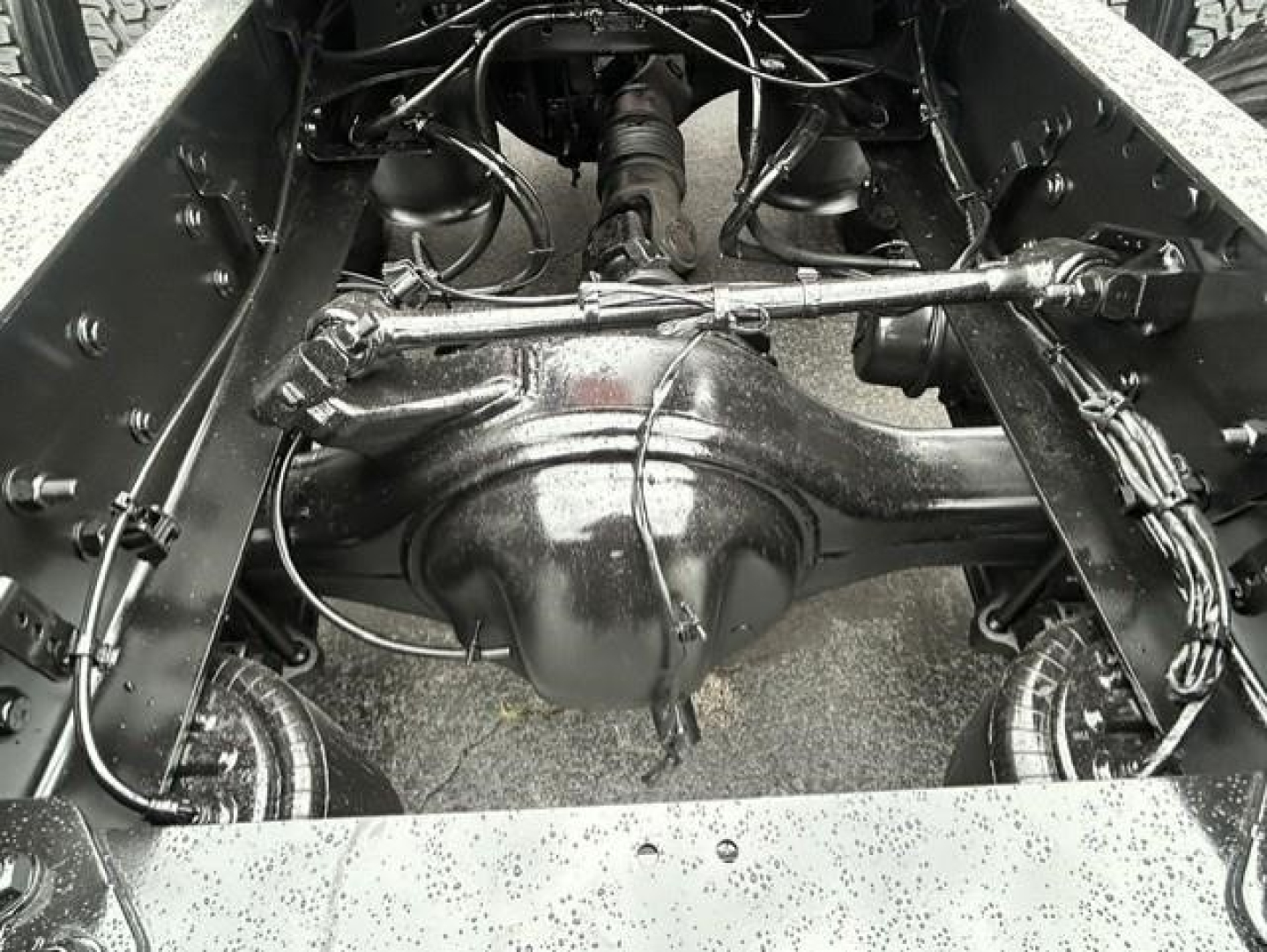 Freightliner Cascadia chassis rear view showing solid steel axle housing and suspension links with wiring harnesses and leaf springs in center; wet road grime on frame.