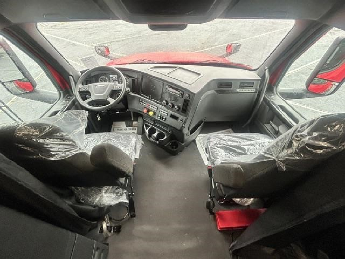 Dashboard and front interior of a 2023 Freightliner Cascadia truck cab with steering wheel, instrument cluster, and driver/passenger seats covered in plastic.