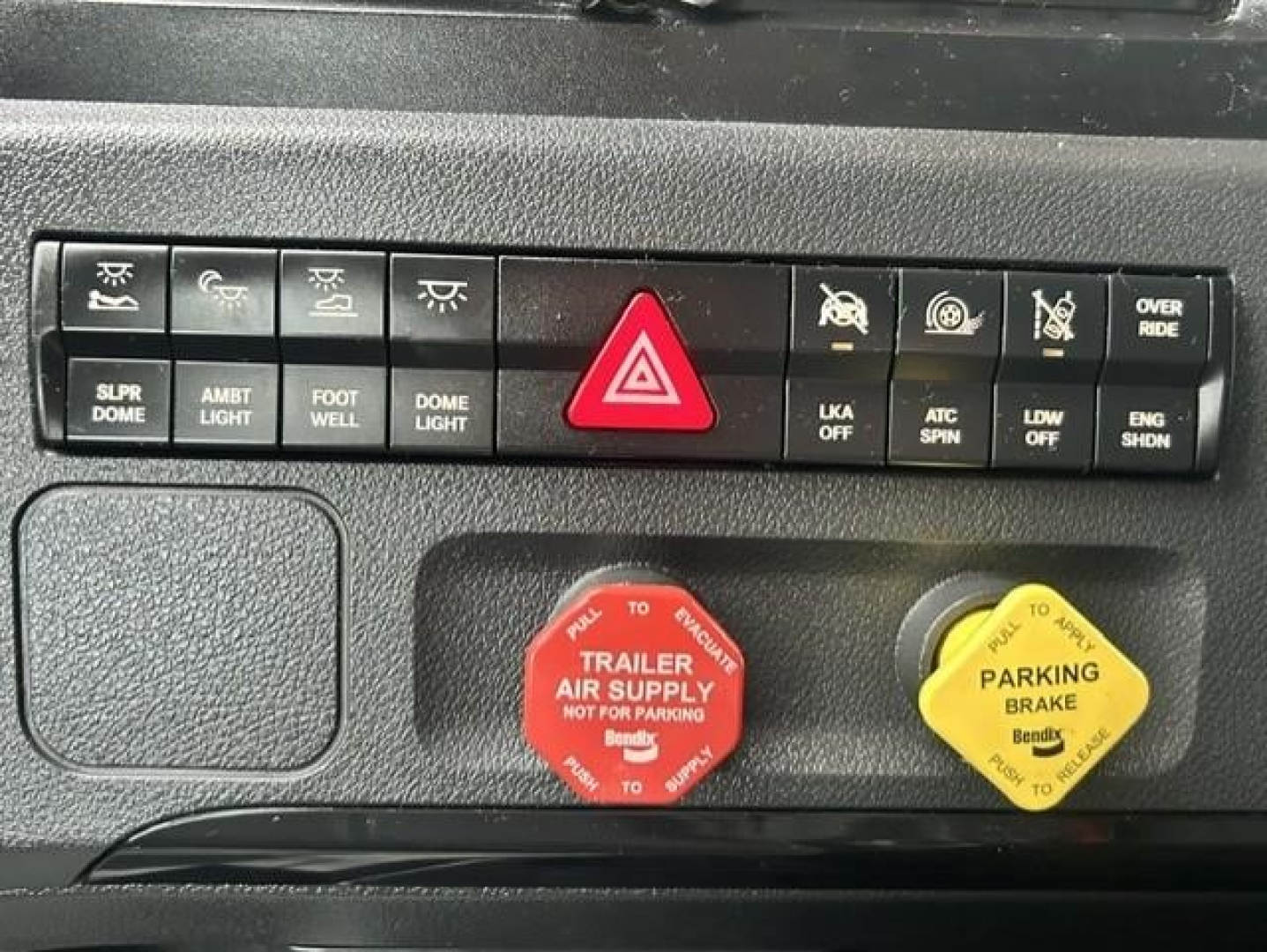 Dashboard controls showing hazard triangle button, various switches labeled: SUP DOME, AMBIENT LIGHT, FOOT WELL, DOME LIGHT, LKA OFF, ATC SPIN, LDW SHDN, ENG SHDN, OVER RIDE, parking brake knob yellow, trailer air supply red knob with text,
