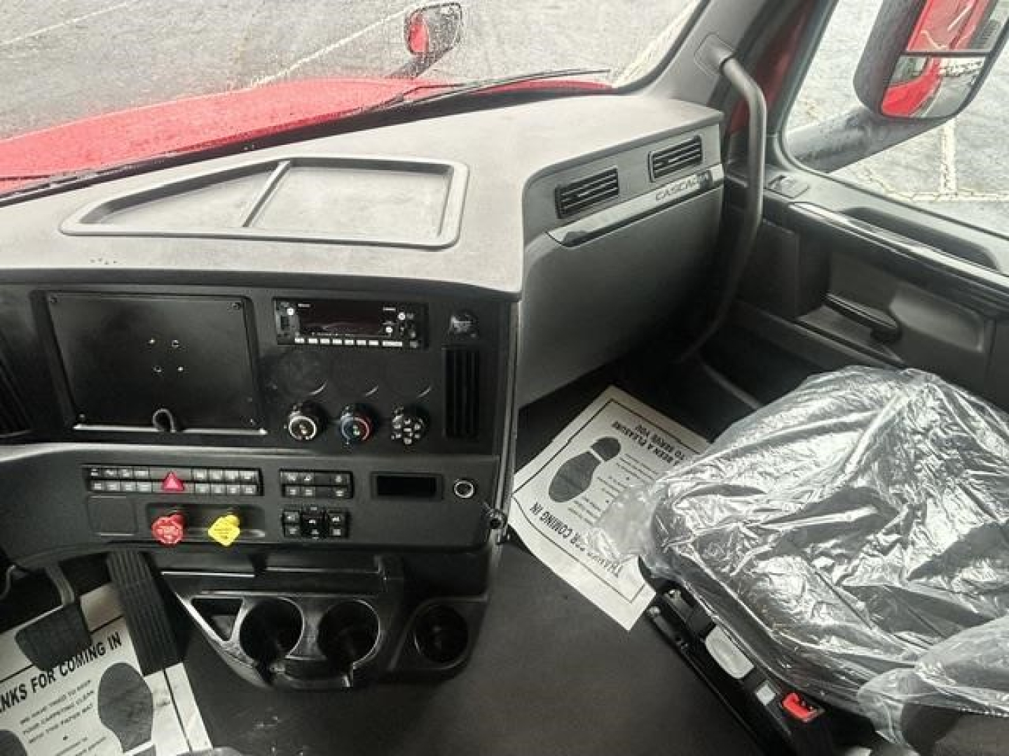 Cab interior of a Freightliner Cascadia truck showing dashboard with controls, steering area, and driver seat covered in plastic.