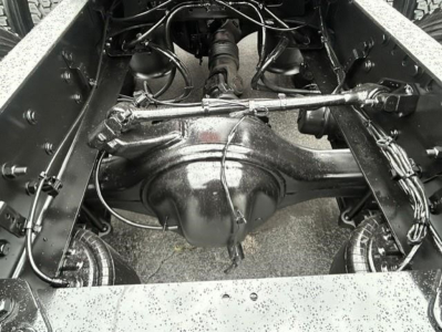 Freightliner Cascadia chassis rear view showing solid steel axle housing and suspension links with wiring harnesses and leaf springs in center; wet road grime on frame.
