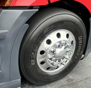 Front left wheel of a Freightliner Cascadia showing Bridgestone tire and chrome hubcap, partial red cab and gray fender.