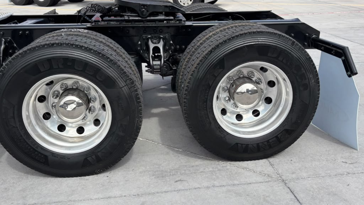 Kenworth T680 truck axle area showing dual rear axles with chrome wheels and tires, undercarriage components, and a partial blue mud flap.