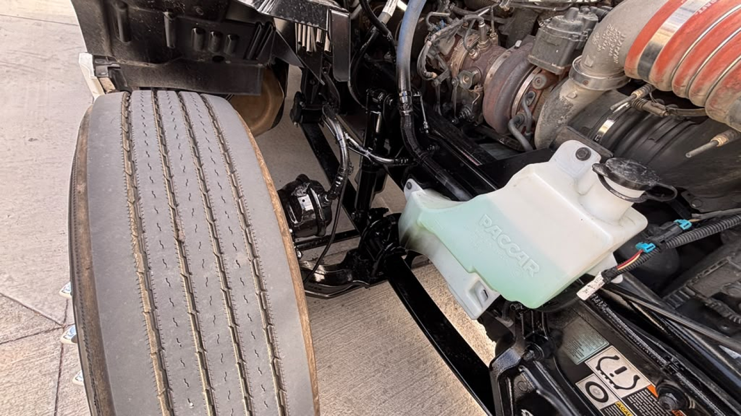 Truck engine bay of a 2023 Kenworth T680 showing coolant reservoir and air intake hoses, front wheel nearby, with visible engine components and hoses.