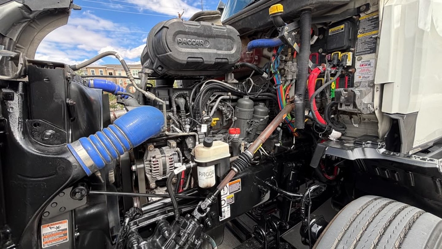 Kenworth T680 engine bay showing engine components, hoses, wiring, and a blue silicone intake hose; various labels and a tank in view.