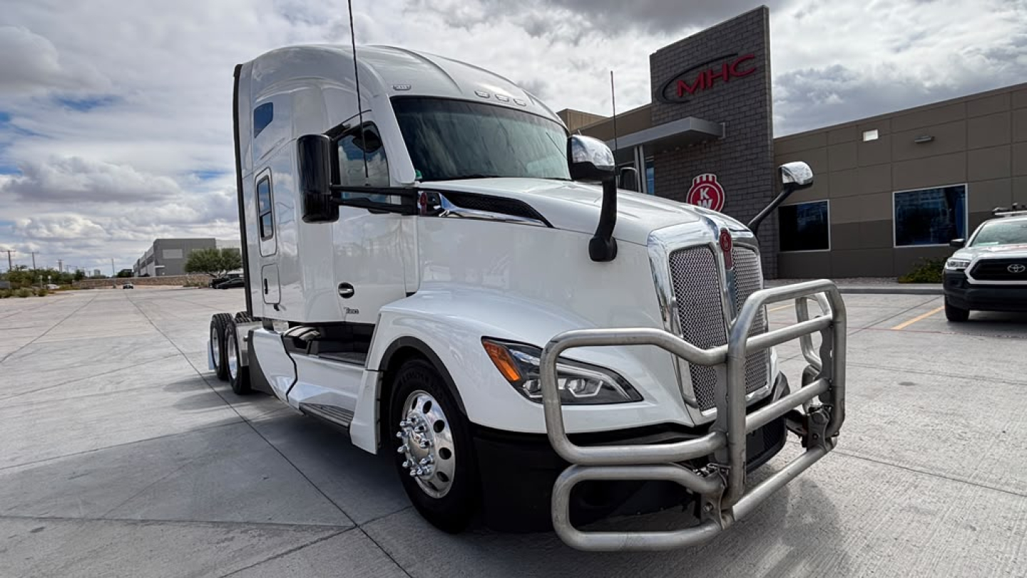 Truck tractor with chrome bumper guard in front, white Kenworth T680 sleeper cab, large grille, side mirrors, and aerodynamic lines; dealership building in background.
