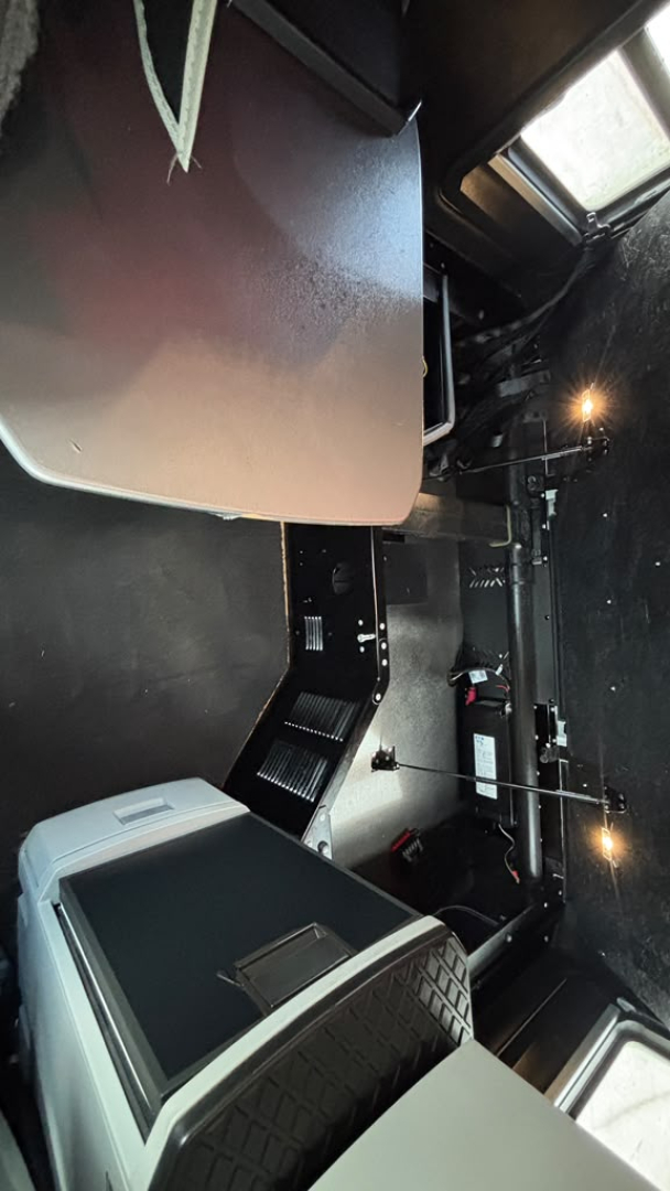 Interior dash area of a 2023 Kenworth T680 showing control panels and storage below the driver seat region; visible components include a textured step panel and a portion of the dashboard.
