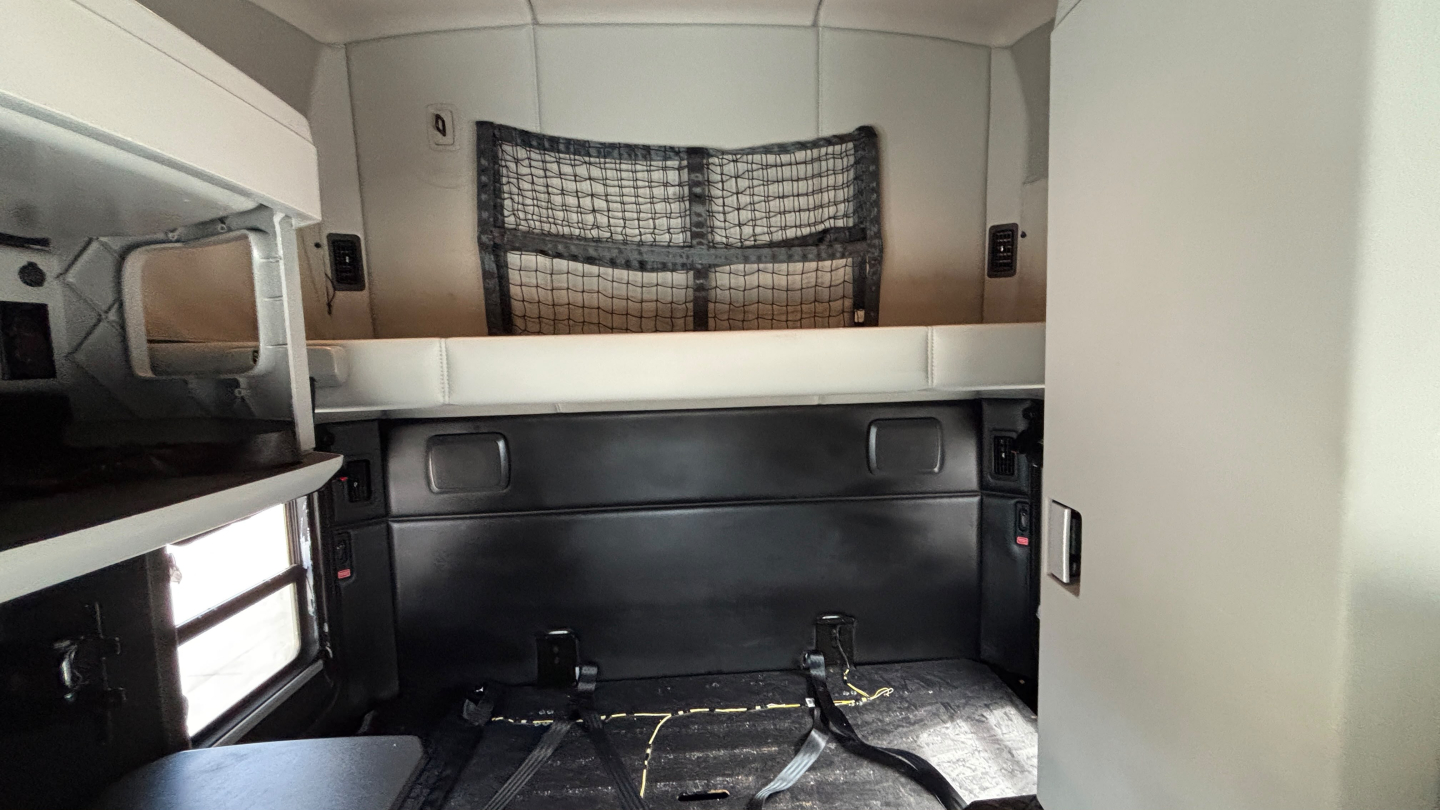 Interior sleeper cab of a Kenworth truck with side storage cabinets and a cargo net at the back wall. No visible branding text beyond the truck's interior features.