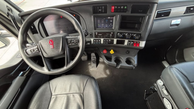 Kenworth T680 cockpit view showing steering wheel, dashboard with center touchscreen, control buttons, gear shifter, and two cup holders; nozzled total visible.
