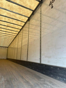 Trailer interior showing white side walls with vertical support rails, wooden floor, and a black scuff guard along the bottom. Small text near top corner reads '40' or '48' with arrows.