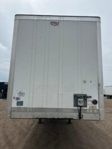 Rear view of a white Wabash trailer with riveted steel panels, bumper lock box, and electrical connections mounted on the lower right side.