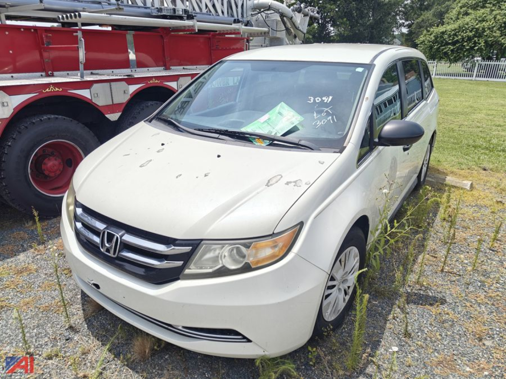 2014 Honda Odyssey minivan in a used lot, light-colored body with front grille and headlights visible, windshield with papers, passenger-side view.