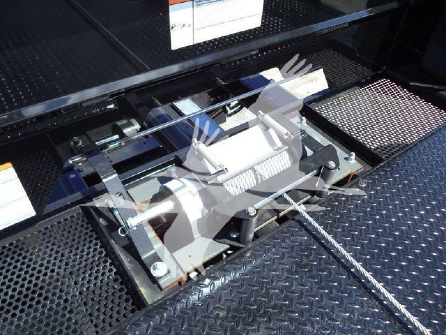 Truck rollback equipment winch assembly mounted in metal diamond plate bed showing winch drum and steel cable.