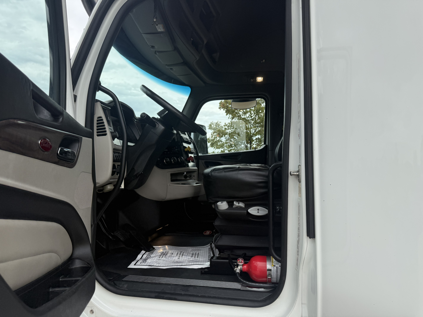 Peterbilt 579 truck interior view of cabin with steering wheel, dashboard, and entry area visible from open driver door; driver seat and floor mats present; red fire extinguisher near the doorway.