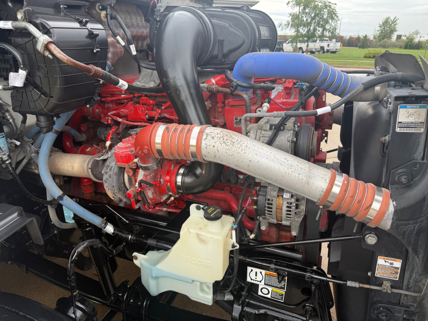Peterbilt 579 engine bay showing red diesel engine, hoses, alternator, coolant reservoir, and air intake components.
