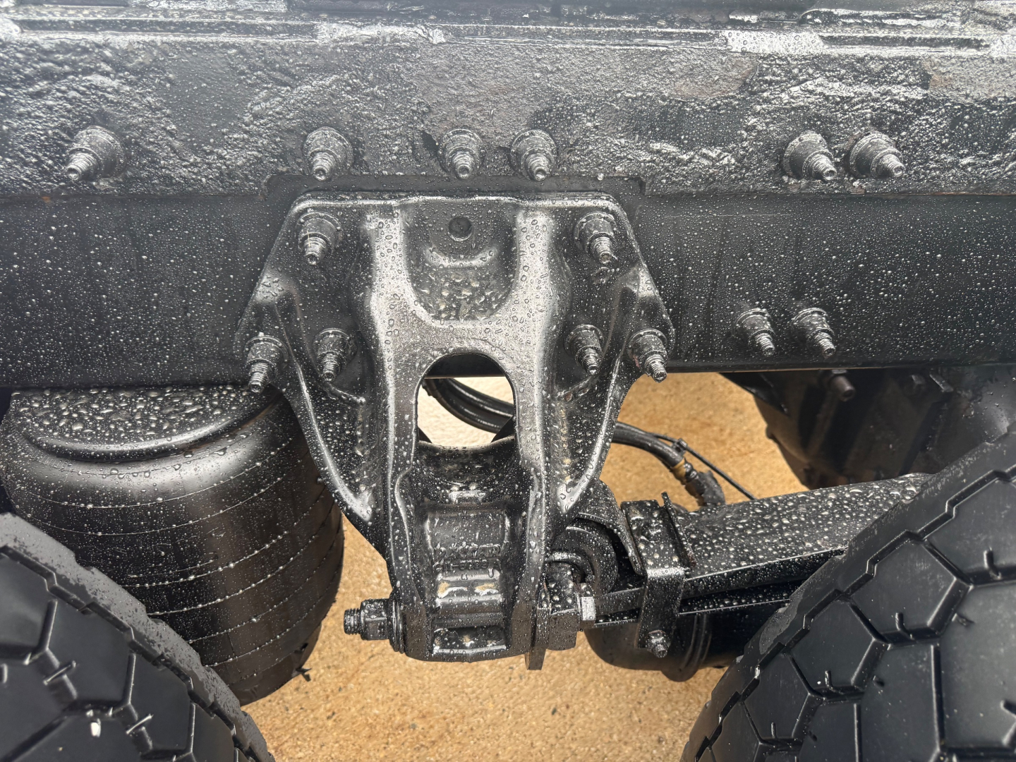 Peterbilt truck chassis view showing rear suspension hanger with multiple bolts and leaf spring, dampened by mud and grime. Partial wheel and tire visible.