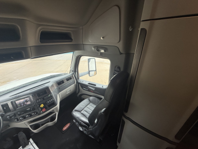 Peterbilt 579 truck interior cab view showing driver seat, steering wheel, dashboard, and overhead storage; side window and door area; part of sleeper area visible.