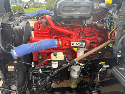 Peterbilt 579 engine bay showing red Cummins X15 engine, blue intercooler hose, coolant reservoir, air intake, and numerous hoses and wiring.