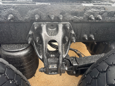 Peterbilt truck chassis view showing rear suspension hanger with multiple bolts and leaf spring, dampened by mud and grime. Partial wheel and tire visible.