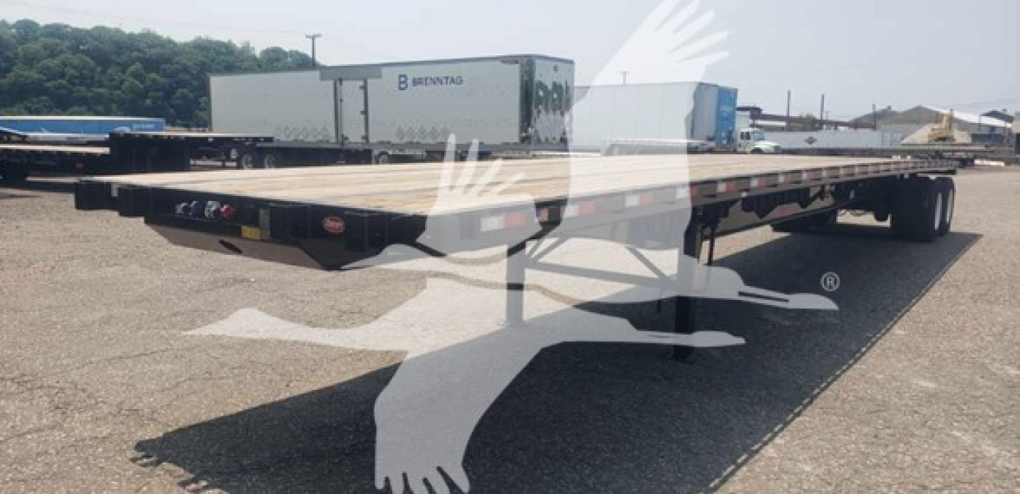 Flatbed trailer in a parking lot showing a long low profile platform with tandem axles and fifth wheel area.