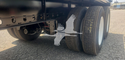 Trailer undercarriage showing tandem axles and dual tires with white rims. Visible are suspension components and a sticker with the bird watermark. No legible model text.