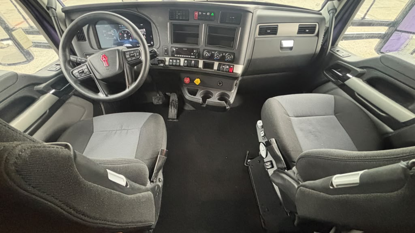 Kenworth T680 truck cab interior showing steering wheel, dashboard, and two front seats. Text on dash includes gauges and controls, with center console and gear shifter visible.