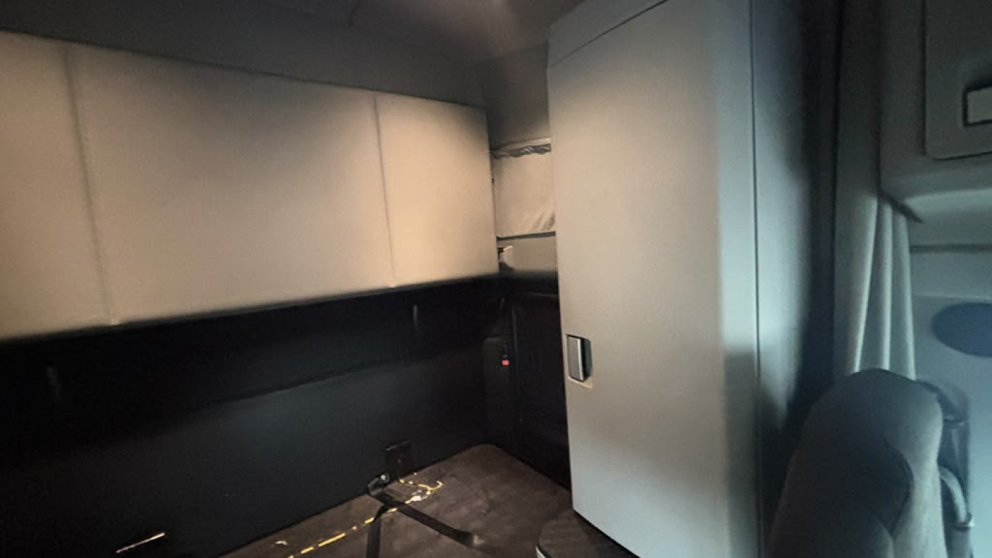 Kenworth T680 cab interior showing sleeper area and cabinet door, with a dark bunk space and light gray storage unit.