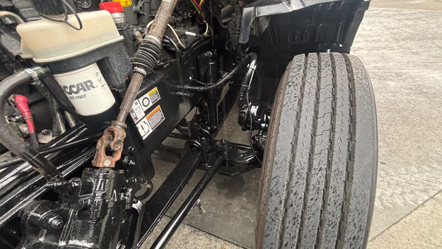 Kenworth T680 engine bay and front suspension view showing brake lines, filter, and wheel. Visible components include coolant reservoir, hoses, and chassis rails.