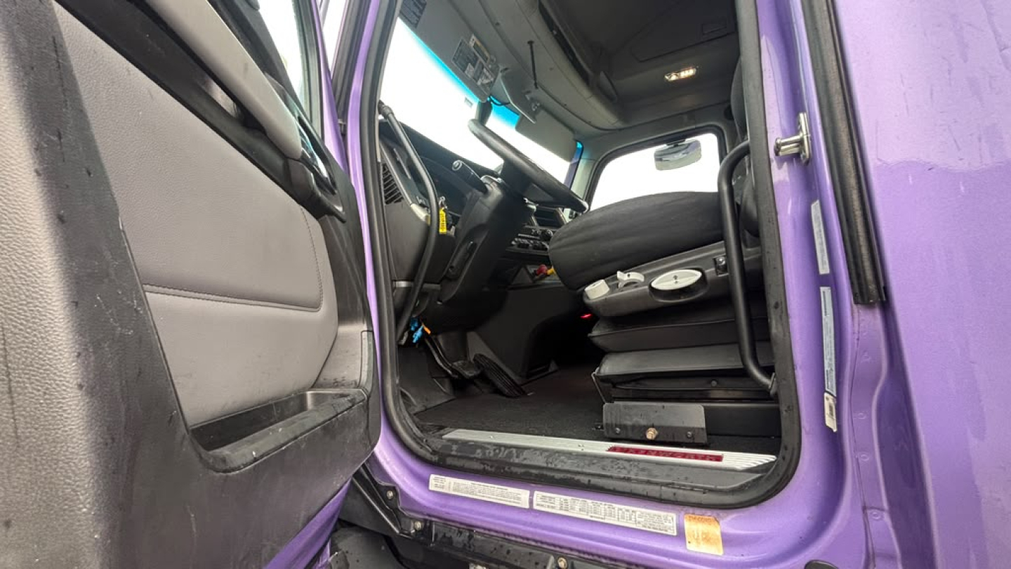 Truck cab interior showing driver's seat and steering wheel of a purple Kenworth T680 with open door.