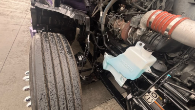 Kenworth T680 engine bay side showing coolant reservoir with blue cap and turbo intake with orange hose, visible hydraulic lines and components.