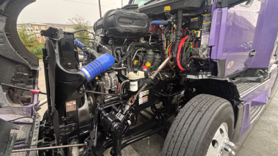 Kenworth T680 engine bay view showing dual air intake, hoses, and visible components in front wheel well; purple truck body in background.