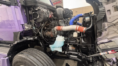 Truck engine bay of a 2024 Kenworth T680 showing turbocharger, hoses, coolant reservoir, and other engine components in a purple-painted cab area.