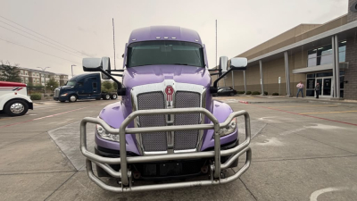 Front of a purple 2024 Kenworth T680 truck with a heavy-duty grille guard in a parking lot.