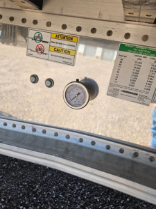 Dashboard area on a Manac trailer showing a pressure gauge and warning labels. Close-up of stickers with caution text and a small placard with load info.