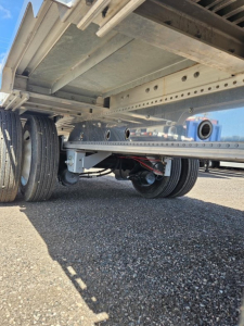 Semi-trailer undercarriage showing tandem axles and suspension with metal drop deck frame and support beams.