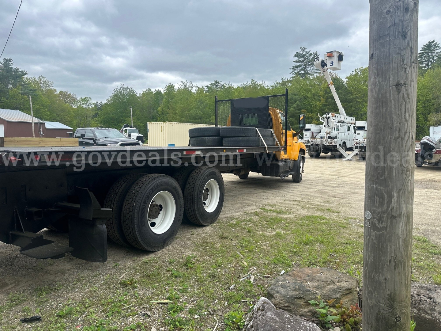 Flatbed truck chassis with dual rear axles and long flatbed. Background shows utility equipment and a bucket lift. Copy says 2006 GMC C8500 with VIN 1GDT8C4C56F417000.