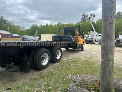 Flatbed truck chassis with dual rear axles and long flatbed. Background shows utility equipment and a bucket lift. Copy says 2006 GMC C8500 with VIN 1GDT8C4C56F417000.