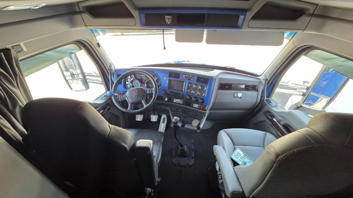 Interior of a Kenworth truck cab, steering wheel on left, dashboard with gauges and digital display, blue accents, seating for driver and passenger.