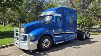 Kenworth T680 tractor in blue with grille guard, semi-truck cab and dual rear axles visible in a parking lot.