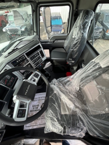 Cab interior of a new 2026 Mack GR64B vacuum tank truck showing steering wheel, dashboard with controls, and plastic-wrapped driver and passenger seats.