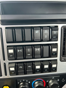 Dashboard switches and controls of a Mack truck cab, including multiple rocker switches with symbols and labels such as PTO, ATC OFF, MUD SNOW, TRLR AUX, and other blank/slotted switches.