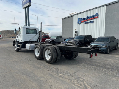 Truck chassis with cab and dual rear axles in a lot outside dealership building.
