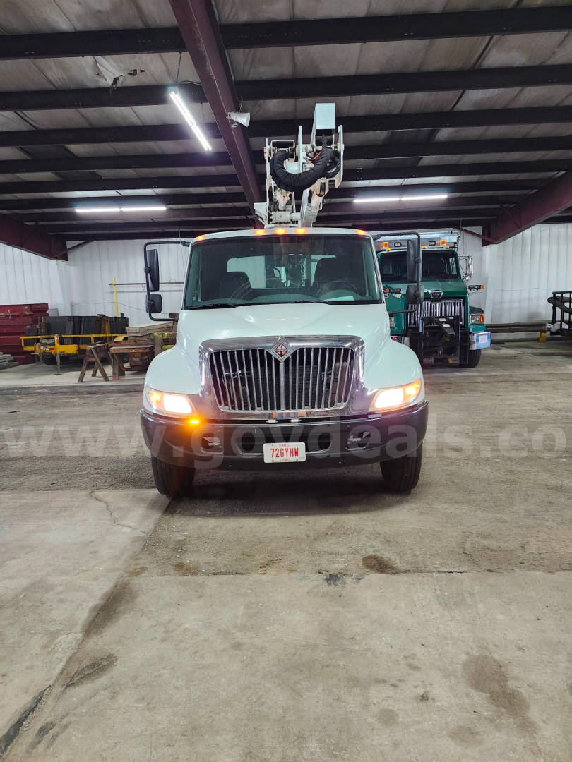 Front view of a white 2004 International 4200 bucket truck with boom raised inside a warehouse, headlights on, stock number 11500-234 visible.