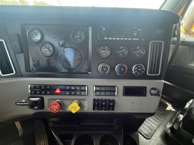 Dashboard panel of a Freightliner Cascadia truck showing gauges, switches, and a black rectangular display; visible text includes TRAILER BRAKE, red hazard button, yellow dial grip, various knobs and vents.