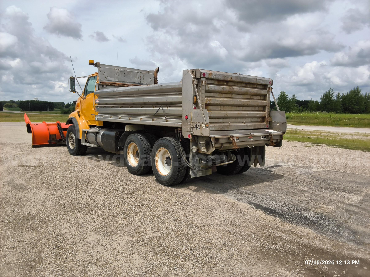 Dump truck from rear-left view showing orange cab, metal dump bed, and twin rear axles with tires; overcast sky.