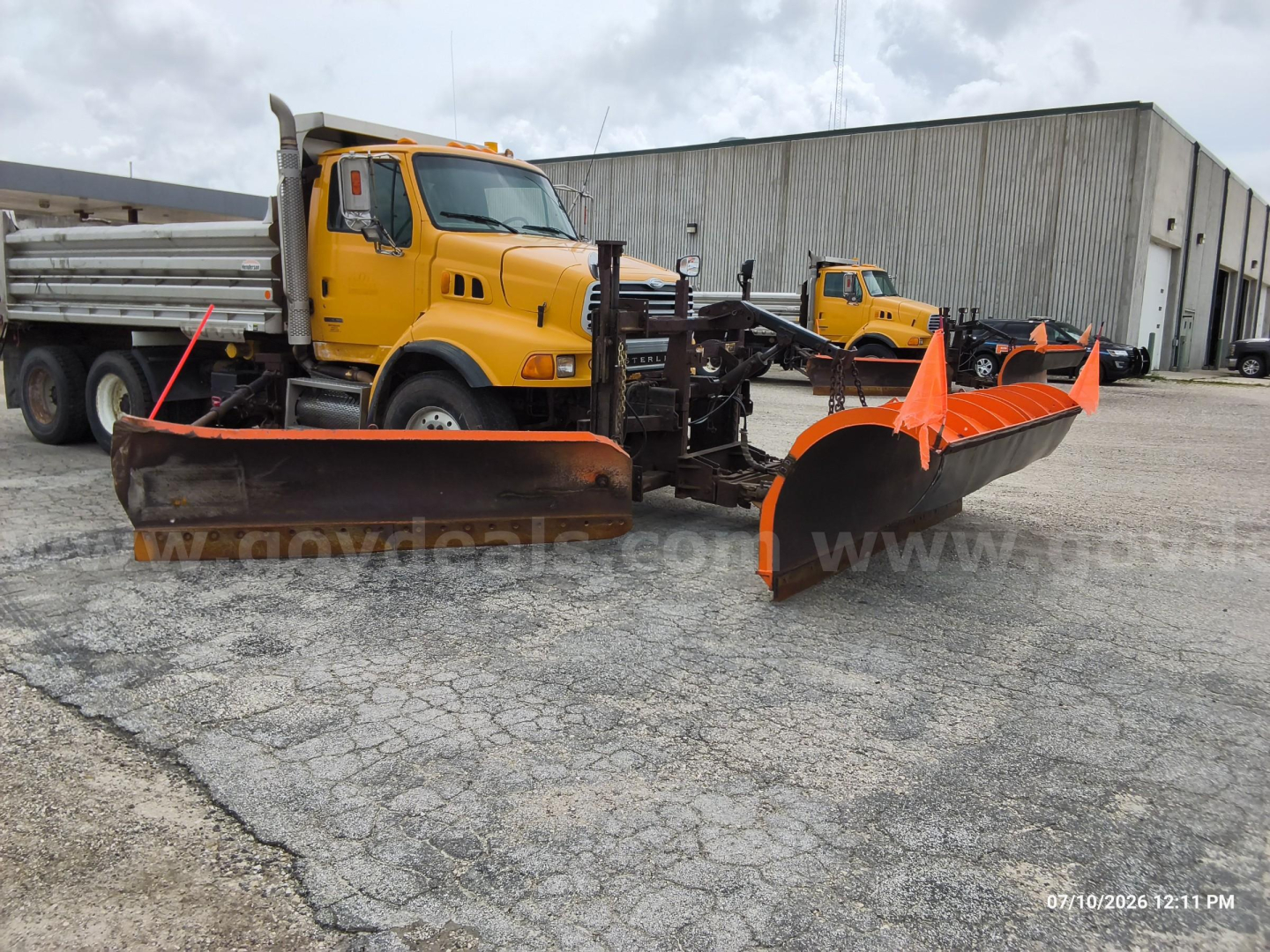 View of a yellow Sterling LT8500 dump truck with a large orange snow-plow blade attached in front. The sticker text on the truck is not fully legible, and there is a warehouse building in the background.