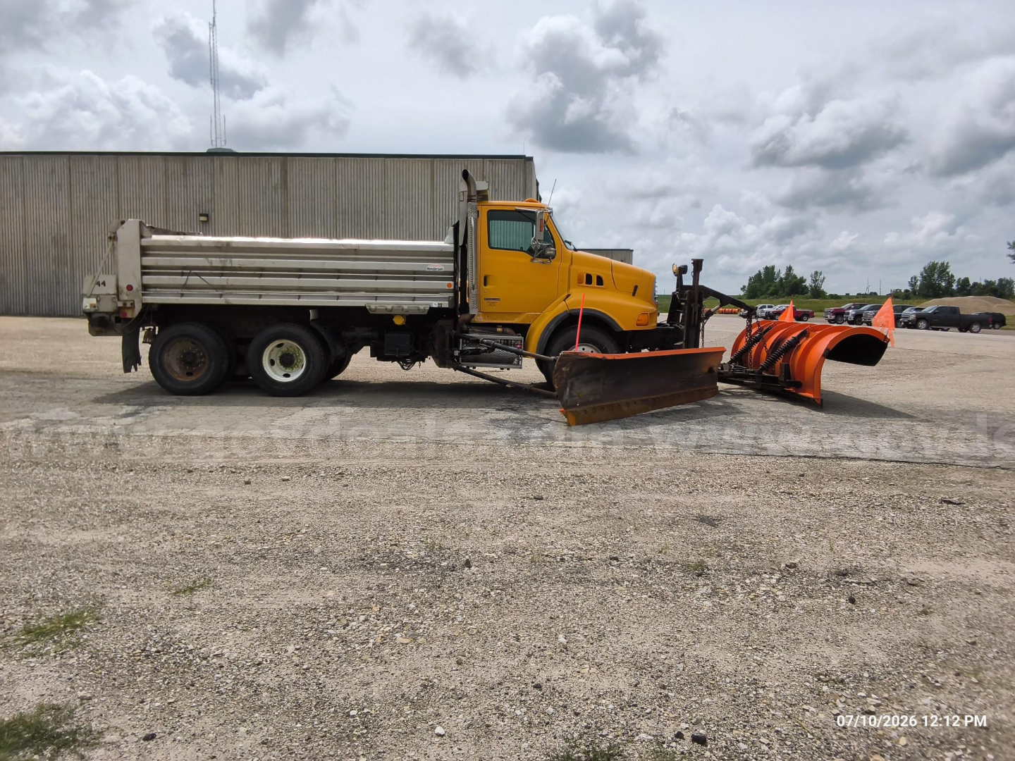 Dump truck with plow and orange snow piper on front, yellow Sterling LT8500 with cargo bed and trailer hitch in an open lot.