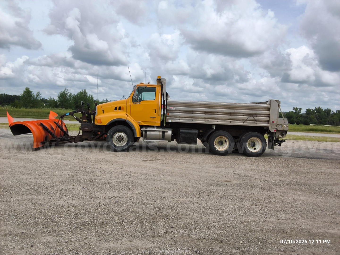 Truck cab and dump bed with attached snow plow or front attachment in front of truck, Sterling LT8500, orange cab, gray dump body, six-wheel configuration, on gravel.