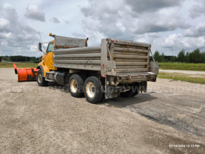 Dump truck from rear-left view showing orange cab, metal dump bed, and twin rear axles with tires; overcast sky.