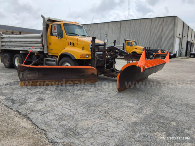 View of a yellow Sterling LT8500 dump truck with a large orange snow-plow blade attached in front. The sticker text on the truck is not fully legible, and there is a warehouse building in the background.